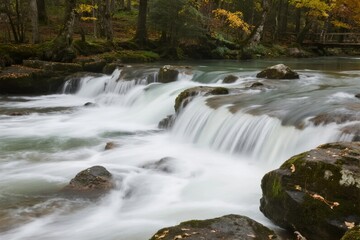 A flowing river cascades over moss-covered rocks in a forested area with autumn foliage.