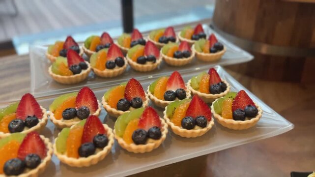 Close up display video of mini fruit tarts with strawberries blueberries and kiwi arranged on a tray, showing fresh ingredients and an inviting dessert atmosphere
