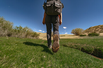 A lone female hiker walks across a rare patch of green grass in the Mongolian desert. Low rear-angle shot captures her boot sole against the backdrop of distant sand dunes.