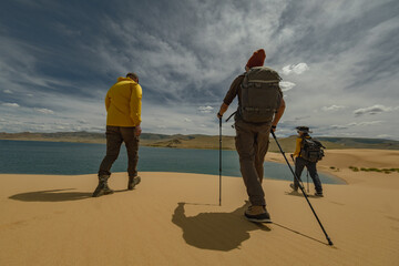 Hikers with trekking poles and backpacks walk along a sand dune overlooking an emerald lake in the desert. Mountain hills and a cloudy blue sky form the backdrop.