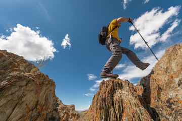 A hiker in bright clothing with poles and a backpack walks along the edge of a rocky mountain. Wide shot from a low angle captures the figure against a blue sky with white clouds.