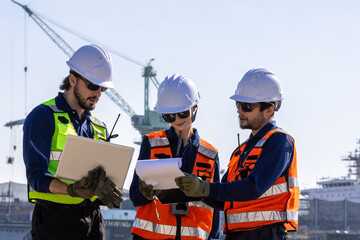 group of industrial engineers workers in a refinery - oil and gas processing equipment and machinery, engineers collaborate with a laptop, blueprint, and digital tablet at the oil storage tanks site.