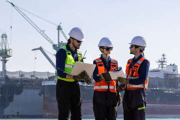 group of industrial engineers workers in a refinery - oil and gas processing equipment and machinery, engineers collaborate with a laptop, blueprint, and digital tablet at the oil storage tanks site.