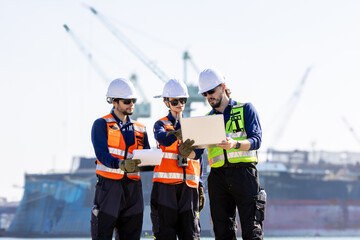 group of industrial engineers workers in a refinery - oil and gas processing equipment and machinery, engineers collaborate with a laptop, blueprint, and digital tablet at the oil storage tanks site.