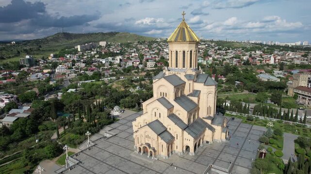 Aerial drone view over the Holy Trinity Cathedral (Sameba) in Tbilisi, Georgia, featuring the iconic golden dome and surrounding cityscape. High-quality 4K footage.