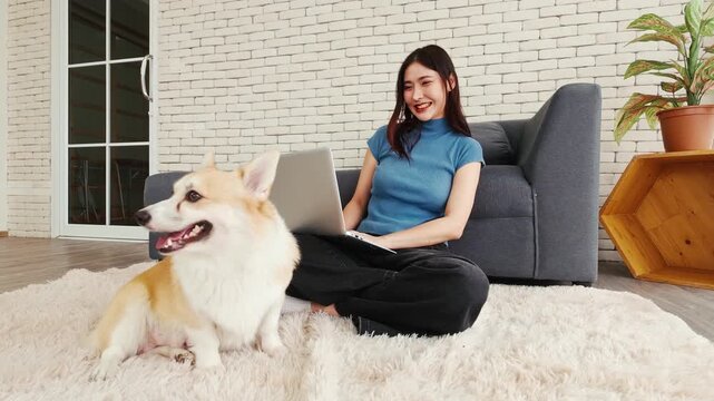 Young woman working on a laptop while lying on a rug beside her Corgi dog at home. A cozy, pet-friendly remote work setup showcasing happiness, productivity, and the comfort of working with pets.
