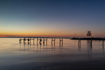 Stiller See bei D&auml;mmerung mit Stegen und Mondsichel, Ein ruhiger See in der Abendd&auml;mmerung mit h&ouml;lzernen Pf&auml;hlen im Wasser. Der Himmel zeigt einen sanften Farbverlauf von warmen Orange- zu k&uuml;hlen Blau