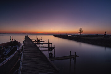 Fototapeta premium Holzsteg am ruhigen See in der blauen Stunde, Ein langer Holzsteg führt über einen stillen See in die Abenddämmerung. Der Himmel zeigt einen sanften Farbverlauf von warmem Orange zu tiefem Blau, währe
