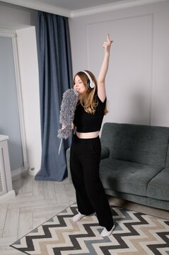 Cheerful young woman of European descent sings into duster while cleaning house and listening to music on headphones. Good mood during leisure time at home.