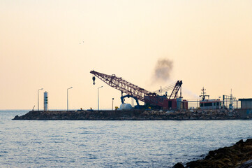 Obraz premium Large red crane with hanging hook operating on rocky pier beside cylindrical structure and docked vessel—framed by calm sea, hazy sky, and active coastal infrastructure with rising smoke.