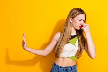 Young woman making a grimacing gesture while holding nose against yellow background, expressing dislike with hand gesturing displeasure