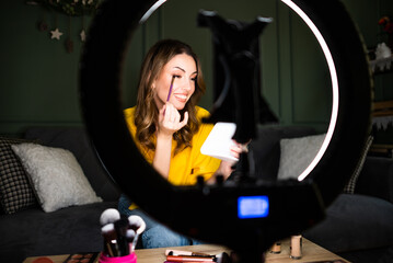 Beauty vlogger filming a makeup tutorial at home. She is sitting on sofa and applying eye shadow.