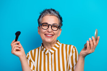 Senior woman in striped shirt with glasses smiling while applying makeup and holding a mirror against a bright blue background for lifestyle and advertising concepts