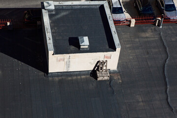 Aerial view of black roof framed by parked cars and a red fence in the background. Flat rooftop features a small central structure with a black roof, pale walls, and a vent on top