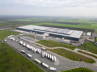 Aerial view of a large logistics warehouse with loading docks and trucks, distribution center surrounded by farmland. Supply chain and transportation concept