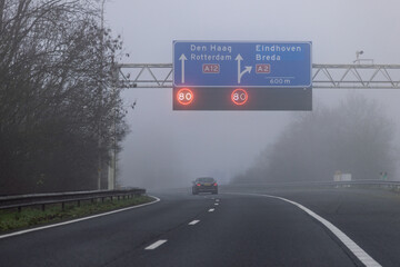 A misty highway scene in the Netherlands with overhead matrix signs displaying an 80 km/h speed limit on the A12 towards Den Haag and Rotterdam.