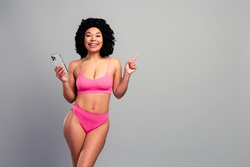 Young confident woman in pink bikini smiles holds phone and points up against gray background for lifestyle fashion and advertising use