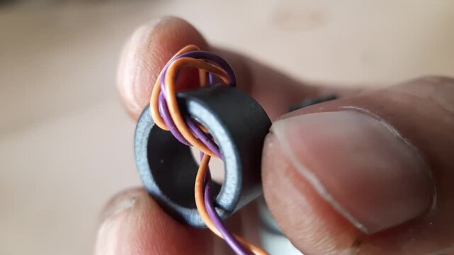 Hand holding a black ferrite toroid core with thin purple and orange copper wire windings in a macro shot