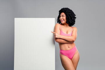 Young woman in pink lingerie smiles and points to blank board in studio