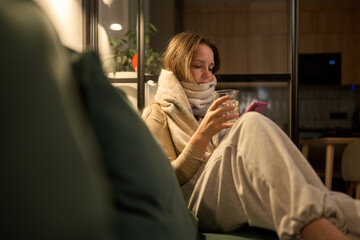 Female wrapped in scarf holding water and phone on sofa indoors