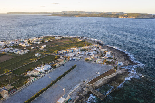 Aerial view of a coastal village where the rugged coastline meets the calm sea, with fields and buildings glowing under the soft light, L-Armier, Il-Mellieha, Malta.