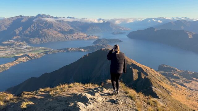A girl walks toward a breathtaking panoramic view at the summit of Roy&rsquo;s Peak in Wanaka, New Zealand. Mountains, lakes, and expansive alpine scenery create a dramatic scene.