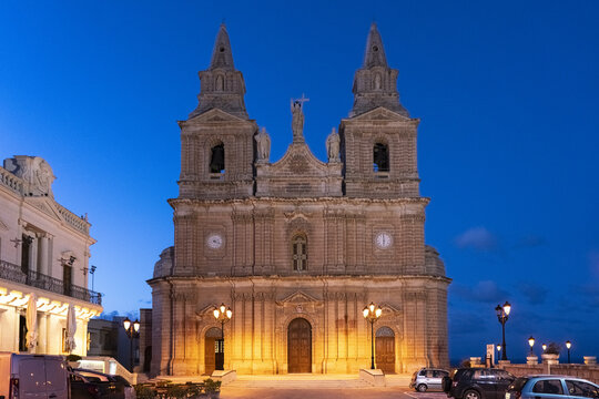 View of the Mellieha Parish Church stands majestically against the deep blue twilight sky, its golden lights casting a warm glow on the stone facade, Mellieha, Malta.