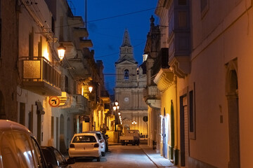 View of golden light bathing ancient stone buildings lining a narrow street leading to a towering church under a deepening blue sky, Mellieha, Malta.