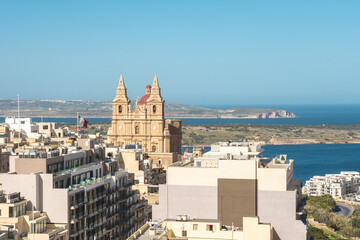 View of Parish Church of Mellieha, its red dome contrasting the clear blue sky, stands amidst a cluster of modern buildings, overlooking the tranquil sea, Mellieha, Malta.