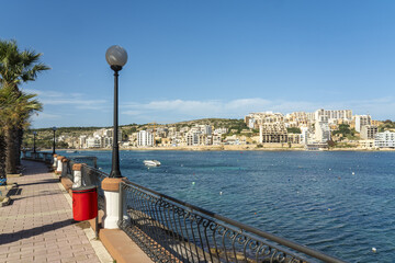 View of a vibrant promenade with palm trees, a red trash can, and a black metal railing overlooking the serene blue waters and city buildings, Mellieha, Malta.