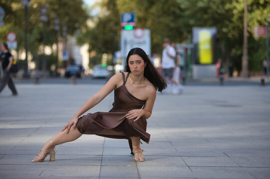 Young woman, beautiful, brunette, blue eyes, Hispanic, wearing an elegant brown chiffon dress and gold heels, posing crouched on a city sidewalk while the wind blows her hair. Concept: model, beauty.
