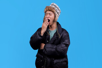 Young man yawning while wearing warm winter jacket and hat against blue background