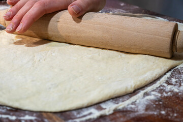 view of hands using a wooden rolling pin to flatten a large sheet of white dough on a dark, flour-dusted surface.
