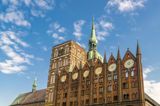View of a grand brick building adorned with clocks and spires reaches towards a sky with fluffy clouds, creating a striking contrast of old and new, Stralsund, Mecklenburg-Vorpommern, Germany.