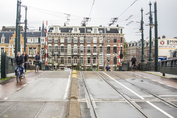 Amsterdam, Netherlands - 17 November 2016: View of cyclists crossing a bridge with tram tracks under a cloudy sky, framed by classic Dutch architecture.