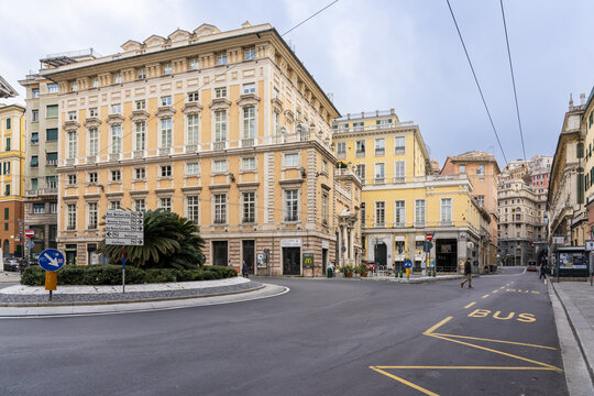 Genoa, Italy - 25 December 2022: View of stately buildings in warm hues framing a roundabout with a splash of green from a central palm, the asphalt reflecting a muted sky.