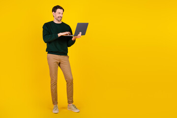 Young man with laptop standing against a bright yellow background wearing green sweater and khaki...