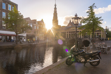 Leiden, Netherlands - 05 May 2019: View of the dark canal water reflecting the sunlit tower and buildings, with a parked motorbike adding a modern touch to the historic scene.