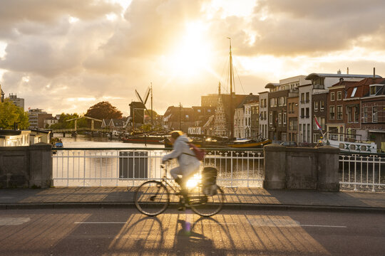 Leiden, Netherlands - 05 May 2019: View of a cyclist crossing a bridge, with warm sunlight illuminating the historic skyline featuring windmills and traditional Dutch architecture.