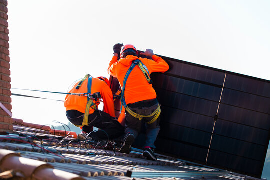 Technicians installing solar panel on roof with harness and helmet outdoors