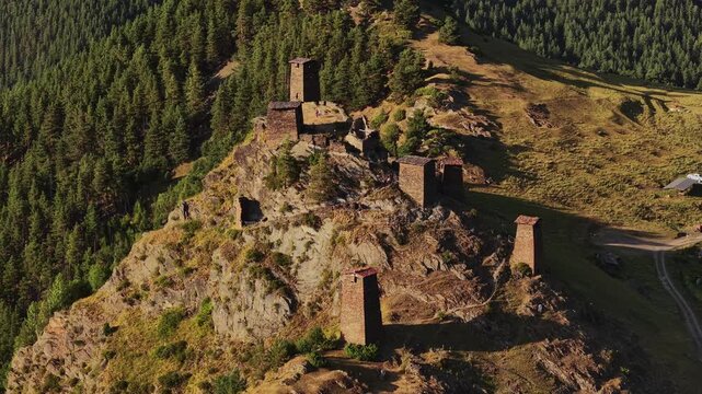 Aerial view of the ancient stone towers of Omalo perched atop a rugged hill, contrasting with the lush green forest and winding dirt road, Omalo, Kakheti, Georgia.