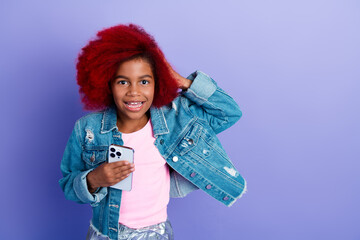 Young girl with pink shirt and denim jacket smiling while holding a phone against a purple...