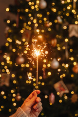 Close-up of sparkler in hand with glowing Christmas tree lights behind