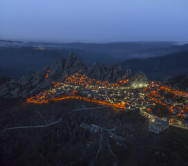 Aerial view of a warm glow embraces the ancient village nestled amidst rugged peaks, lights twinkling like stars against the twilight sky, Pietrapertosa, Basilicata, Italy.