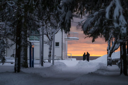 View of a tranquil winter scene with silhouetted figures watching the sunset over the Baltic Sea framed by snow-laden trees and a white building, Parnu, Estonia.