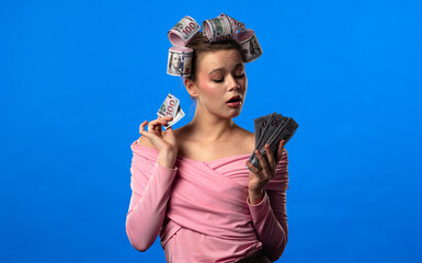 A woman in a pink blouse holding and counting a fan of money, with curlers in the form of banknotes in her hair, against a blue background. A humorous concept of wealth.