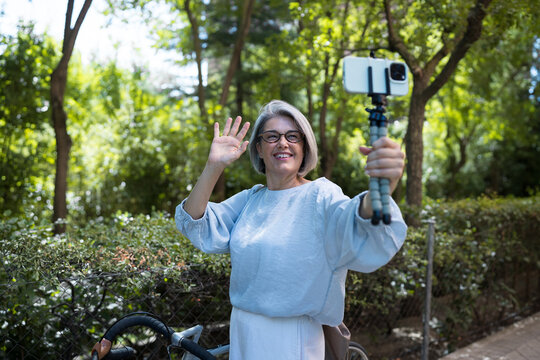 Smiling woman waving while vlogging with phone outdoors near bike