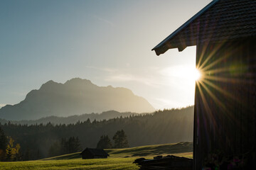 View of the sun shining over the mountain range and green fields, with a wooden structure to the right, creating a striking contrast, Krun, Bavaria, Germany.