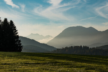 View of a hazy mountain range rising behind a field and dark green trees under a bright sky with cirrus clouds, Krun, Bavaria, Germany.