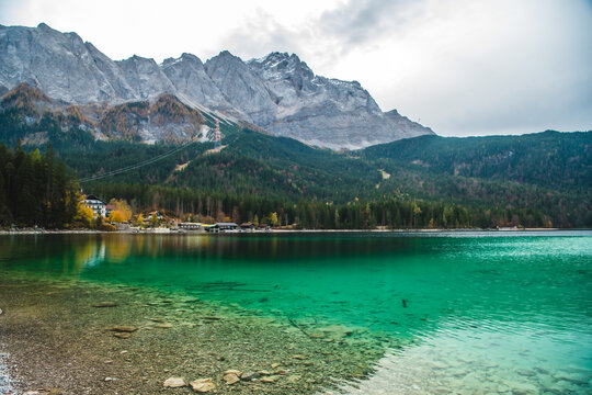 View of the turquoise Eibsee reflecting the dramatic, snow-capped Zugspitze mountains and dense evergreen forests under a cloudy sky, Garmisch-Partenkirchen, Bavaria, Germany.